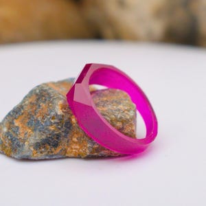 May include: A vibrant, hot pink ring with a geometric design sits on a textured, multi-coloured rock. The ring's facets catch the light, giving it a translucent appearance. The background is blurred.