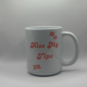 May include: White ceramic mug with the text "Kiss My Tips" in coral-colored retro font, along with coral-colored lip prints and "xox" symbols. The mug has a large handle.