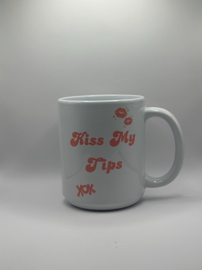 May include: White ceramic mug with the text "Kiss My Tips" in coral-colored retro font, along with coral-colored lip prints and "xox" symbols. The mug has a large handle.