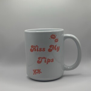 May include: White ceramic mug with a curved handle. The mug features the text "Kiss My Tips" in coral-colored lettering, along with coral-colored lip prints and the letters "xox".