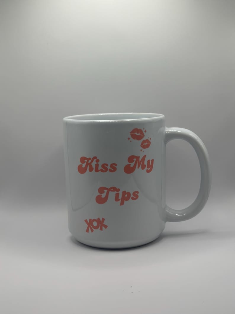 May include: White ceramic mug with a curved handle. The mug features the text "Kiss My Tips" in coral-colored lettering, along with coral-colored lip prints and the letters "xox".