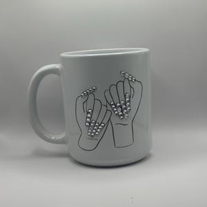 May include: White ceramic mug featuring a black outline illustration of two hands making a pinky promise. The fingernails are adorned with sparkling rhinestones. The mug has a curved handle and a smooth, glossy finish.