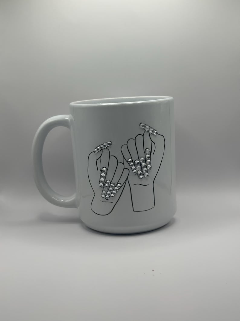 May include: White ceramic mug featuring a black outline illustration of two hands making a pinky promise. The fingernails are adorned with sparkling rhinestones. The mug has a curved handle and a smooth, glossy finish.