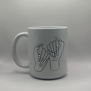 May include: White ceramic mug featuring a black outline illustration of two hands interlocked at the pinky fingers, each with long, embellished fingernails. The nails are adorned with silver-colored rhinestones. The mug has a large handle.