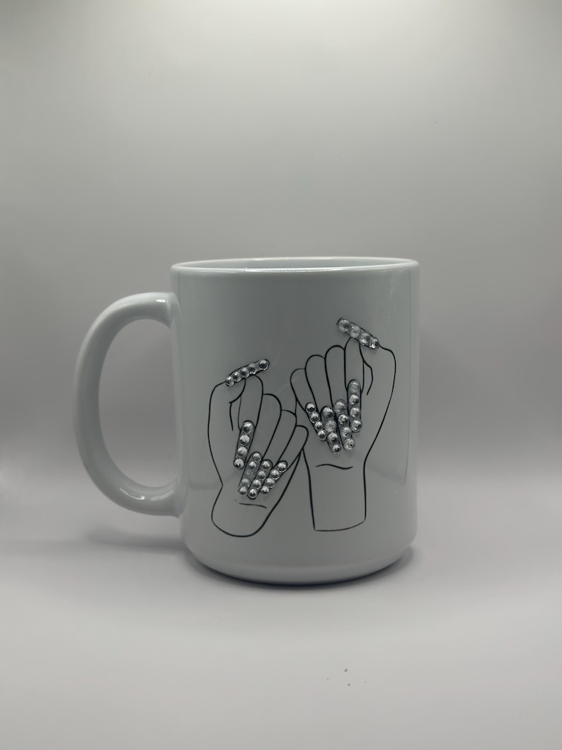 May include: White ceramic mug featuring a black outline illustration of two hands interlocked at the pinky fingers, each with long, embellished fingernails. The nails are adorned with silver-colored rhinestones. The mug has a large handle.