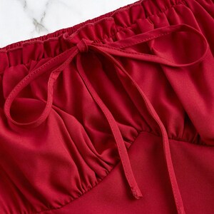 May include: Close-up of a red top with a gathered neckline and a bow detail. The fabric appears to be a smooth, satin-like material. The top is a solid, vibrant red color.