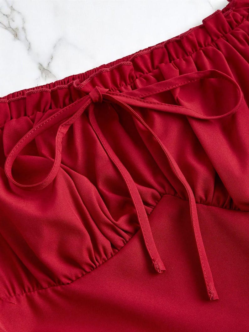 May include: Close-up of a red top with a gathered neckline and a bow detail. The fabric appears to be a smooth, satin-like material. The top is a solid, vibrant red color.
