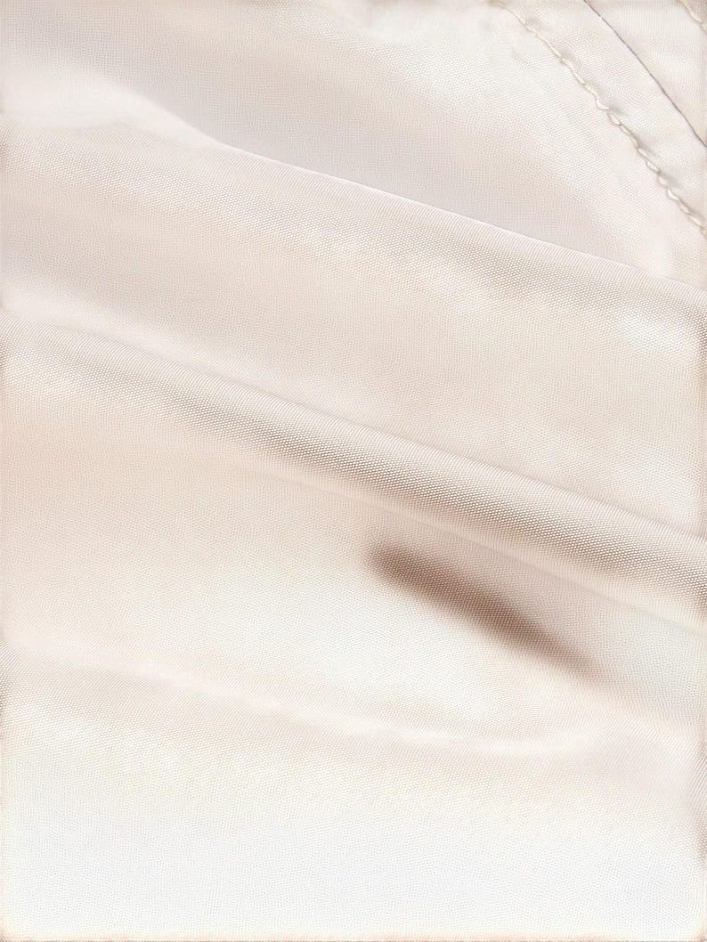 May include: Close-up of a cream-colored fabric with soft folds and subtle shadows. The fabric appears to be smooth and possibly satin or silk, with a slight sheen. The image highlights the texture and drape of the material.