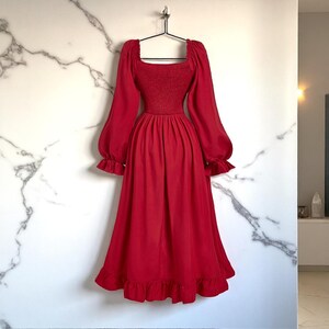 May include: A red dress with a square neckline, long sleeves, and a ruffled hem. The dress has a fitted bodice and a flowing skirt. The sleeves are long and puffy, with ruffled cuffs. The dress is hanging on a black hanger against a white marble wall.