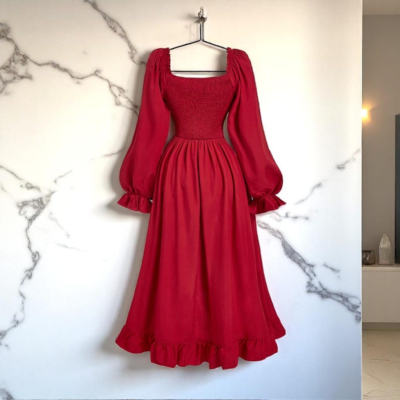 May include: A red dress with a square neckline, long sleeves, and a ruffled hem. The dress has a fitted bodice and a flowing skirt. The sleeves are long and puffy, with ruffled cuffs. The dress is hanging on a black hanger against a white marble wall.
