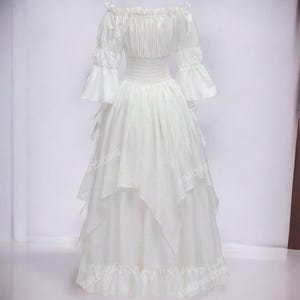 K&ouml;nnte beinhalten: Ein wei&szlig;es, schulterfreies Kleid mit einem mehrlagigen Rock und R&uuml;schenbesatz. Das Kleid hat ein tailliertes Oberteil und 3/4-&Auml;rmel mit R&uuml;schen.