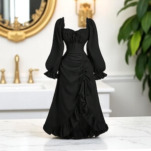 May include: A black dress with long, puffed sleeves and a square neckline. The dress features a ruched bodice and a ruffled hemline. The dress is displayed on a white surface, with a gold-framed mirror and a plant in the background.