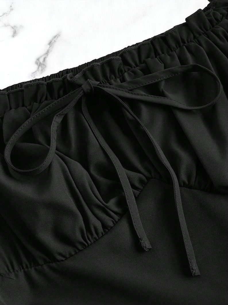 May include: Close-up of a black top with a gathered neckline and a drawstring tie. The fabric is smooth and drapes softly, with subtle folds adding texture. The drawstring is tied in a bow, creating a delicate detail.