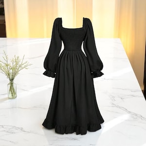 May include: A black midi dress with a square neckline and long, puffed sleeves. The dress features a fitted bodice and a flowing skirt with a ruffled hem. The dress is displayed on a white marble surface.
