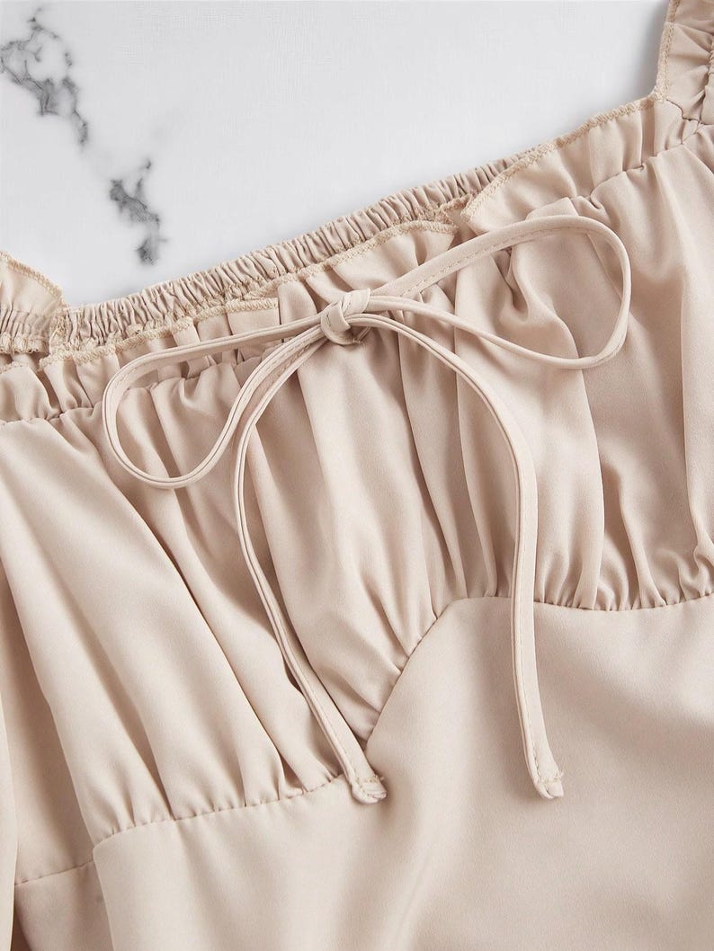May include: Close-up of a beige top with a square neckline and a tie detail. The top features ruffled edges along the neckline and a gathered bodice. The fabric has a smooth, silky appearance.