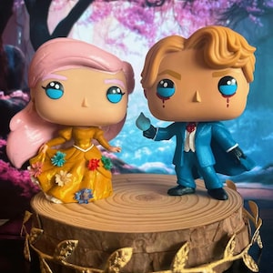 May include: Two Funko Pop figures, one with pink hair in a yellow dress with floral accents, and the other in a blue suit, standing on a wooden base with gold leaf accents. The background features a blurred pink and purple floral scene.