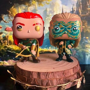Pode incluir: Duas figuras Funko Pop de personagens de fantasia. Uma tem cabelo ruivo, uma roupa verde e dourada e um arco e flecha. A outra tem cabelo loiro e uma máscara e roupa turquesa e dourada. Elas estão em uma base de fatia de madeira com detalhes dourados.