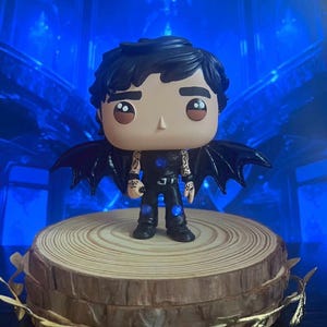 Pode incluir: Uma figura de vinil Funko Pop de um personagem com cabelo preto, asas pretas e uma roupa preta. A figura está em uma base de fatia de madeira com um detalhe decorativo dourado. O fundo é azul desfocado.