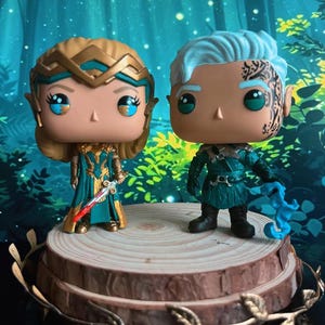 Puede incluir: Dos figuras Funko Pop de personajes de fantasía. Una figura tiene cabello rubio y un atuendo turquesa y dorado, sosteniendo una espada. La otra figura tiene cabello azul y un atuendo turquesa, sosteniendo un objeto azul. Se exhiben en una plataforma de madera.