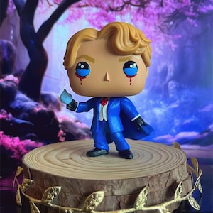 May include: A Funko Pop figure of a character in a blue suit with a cape, holding a blue object. The figure has blonde hair, blue eyes, and red tear streaks. It stands on a wooden base with gold leaf accents.