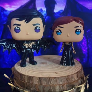 Pode incluir: Duas figuras Funko Pop de personagens de fantasia. Uma figura é um personagem masculino em uma roupa preta com asas de morcego. A outra é uma personagem feminina em um vestido preto. Elas são exibidas em uma fatia de madeira.