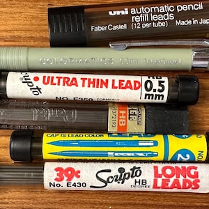 May include: A collection of writing supplies, including a light green mechanical pencil, lead refills, and a container of Uni automatic pencil refill leads. The lead refills are in various sizes and colors, with text labels.