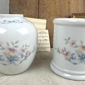 May include: A white ceramic vase with a rounded body and a matching white ceramic cylindrical container. Both pieces feature a delicate floral pattern of pink flowers, green leaves, and light blue ribbons painted around the center of each item.