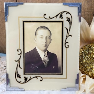 May include: A rectangular glass picture frame featuring a cream-colored border with black filigree corner accents and metallic silver corner clips. The frame holds a black and white portrait of a person wearing a suit and a checkered tie.