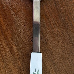 May include: A vintage serving fork with a stainless steel head and a white handle. The handle is decorated with pink flowers and green leaves. The fork is designed for serving food.