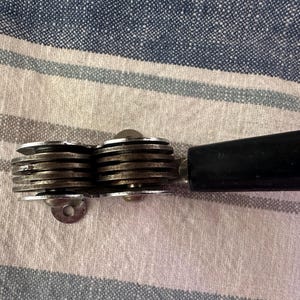 Puede incluir: Un abrelatas vintage con un mango negro y un mecanismo de corte plateado de varias ruedas. El abrelatas est&aacute; sobre un fondo de tela a rayas.