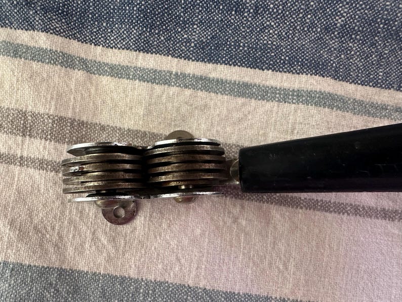 Puede incluir: Un abrelatas vintage con un mango negro y un mecanismo de corte plateado de varias ruedas. El abrelatas est&aacute; sobre un fondo de tela a rayas.
