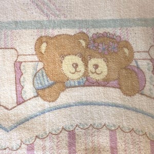 May include: A fabric textile featuring a repeating pattern of two brown teddy bears resting in a bed. The bears are depicted with light brown muzzles and one wears a purple floral crown. The background includes soft pastel stripes in pink and blue.