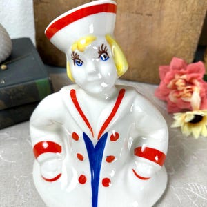 May include: A white ceramic figurine of a person wearing a sailor uniform with a red and blue collar, red sleeve bands, and red buttons. The figure has yellow hair, blue eyes, and a white sailor hat with a red stripe, all in a glossy finish.
