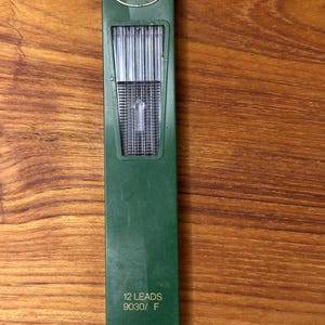 May include: A green Faber-Castell pencil lead container. The container has the Faber-Castell logo at the top and the text "12 LEADS 9030/ F" at the bottom. The container is rectangular and contains pencil leads.