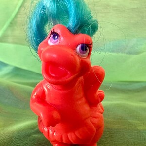 May include: A vintage, red plastic toy duck with teal hair and purple eyes. The duck has a wide open mouth and small wings. The toy is standing on a green surface.