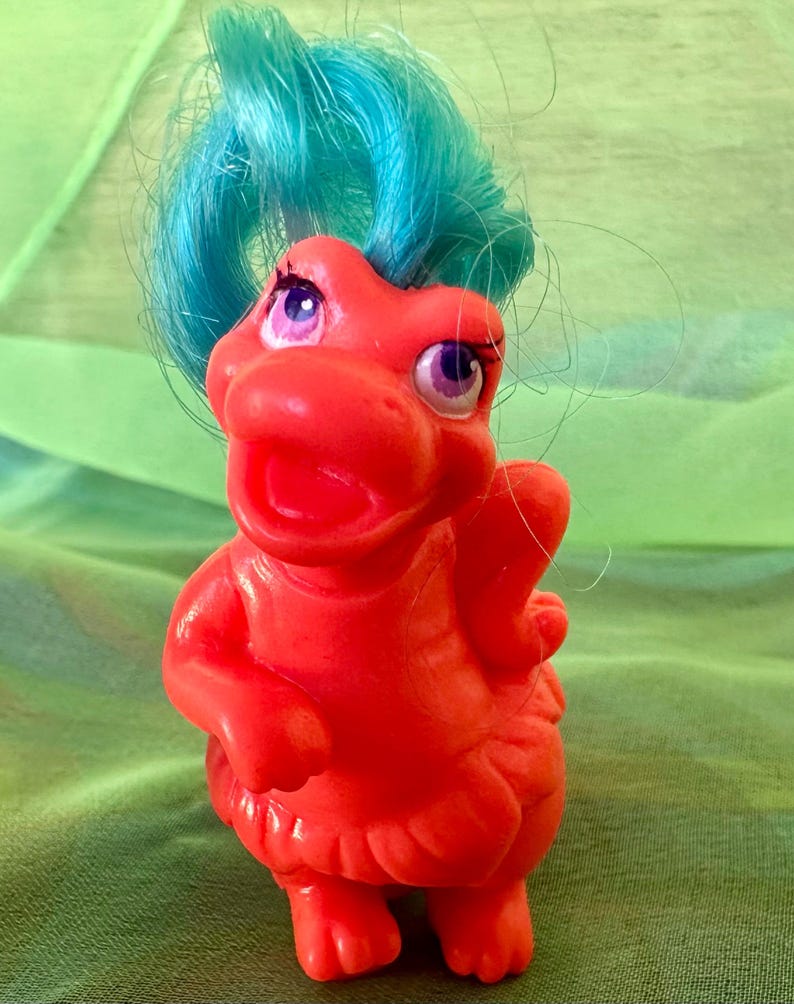May include: A vintage, red plastic toy duck with teal hair and purple eyes. The duck has a wide open mouth and small wings. The toy is standing on a green surface.