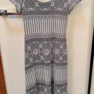 May include: A gray and white patterned dress with short sleeves hangs on a hanger. The dress features a scoop neckline and a detailed design with floral and striped patterns. The dress is made of a lightweight material.