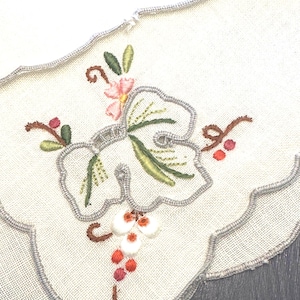 Może przedstawiać: Biały, haftowany bieżnik na stół z falistym brzegiem. Haft przedstawia wzór winorośli z zielonymi liśćmi, różowymi kwiatami i czerwonymi jagodami. Krawędź jest obszyta srebrno-szarą nicią.