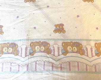 Op de afbeelding: Een witte stof met een herhalend patroon van bruine teddyberen met paarse bloemaccenten. Het ontwerp bevat een horizontale rand met beren die op een geschulpte richel rusten, geaccentueerd door lichtblauwe en paarse strepen en stippen.