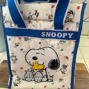 May include: A white and blue Snoopy lunch bag with blue handles and trim. The bag features a large Snoopy graphic and smaller Snoopy illustrations with paw prints and yellow accents. The word "SNOOPY" is displayed across the front.