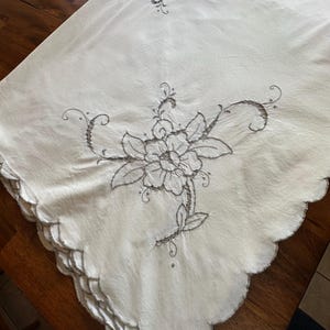 May include: Cream-colored tablecloth with embroidered floral designs. The embroidery features a large central flower and smaller floral accents. The edges are scalloped, adding a decorative touch to the vintage textile.