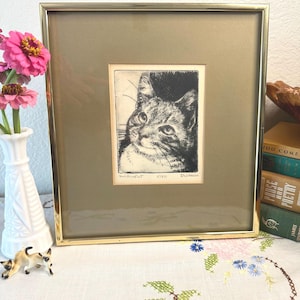 May include: A framed black and white etching of a cat's face, matted in gold and encased in a thin gold metal frame. The print features the handwritten text "Smiling Cat", "5/50", and "Dickson" at the bottom edge of the central image area.