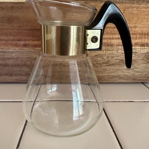 May include: A clear glass coffee pot with a gold band around the middle and a black plastic handle. The pot is sitting on a white tiled surface.