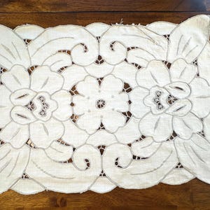 Può includere: Un centrino rettangolare in lino bianco con un complesso ricamo a intaglio. Il design presenta grandi motivi floreali e disegni di foglie a volute con delicati dettagli di cucitura, creando un effetto traforato su tutta la superficie.