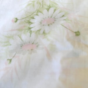Puede incluir: Primer plano de una tela blanca con un estampado floral de margaritas. El diseño presenta grandes margaritas blancas con centros rosas y hojas verdes. El fondo es de un color blanco roto suave, creando una sensación delicada y aireada.