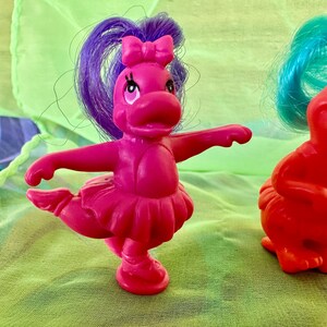 May include: A magenta-colored toy figure of a dancing hippopotamus with a purple ponytail and a pink bow. The hippo is wearing a pink tutu and has its arms outstretched. Another red hippo figure is partially visible in the background.
