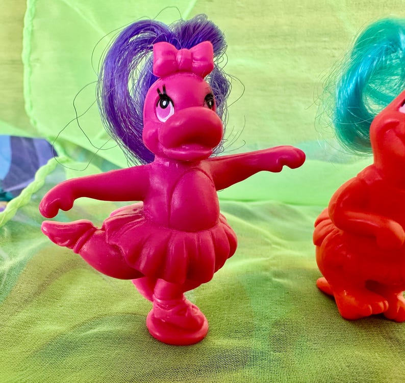 May include: A magenta-colored toy figure of a dancing hippopotamus with a purple ponytail and a pink bow. The hippo is wearing a pink tutu and has its arms outstretched. Another red hippo figure is partially visible in the background.