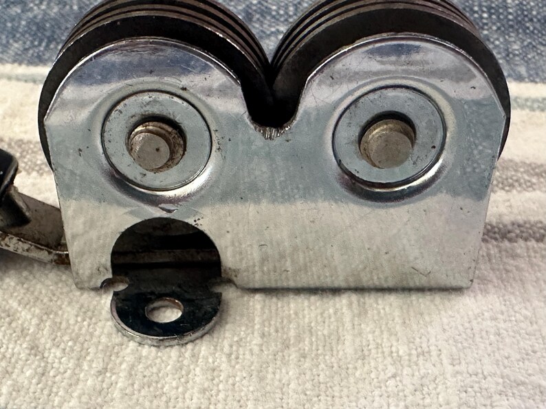 Puede incluir: Primer plano de un afilador de cuchillos manual. El afilador tiene un cuerpo de metal plateado con dos muelas y un mango negro. Las muelas est&aacute;n hechas de un material oscuro y est&aacute;n dise&ntilde;adas para afilar cuchillos.