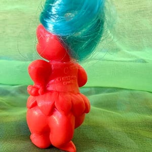 May include: A vintage red Troll doll with bright blue hair. The doll has a round body, a petal-shaped skirt, and a small heart-shaped detail on its back. The doll is made of plastic and has a cheerful expression. The year 1992 is printed on the back.