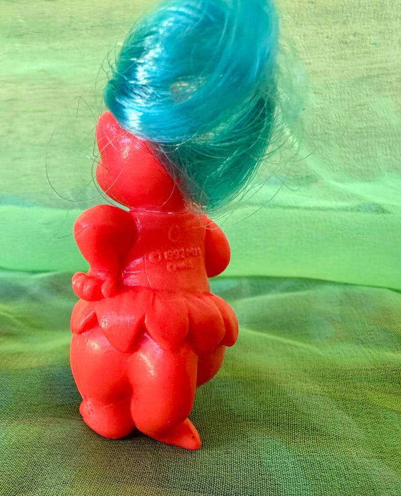 May include: A vintage red Troll doll with bright blue hair. The doll has a round body, a petal-shaped skirt, and a small heart-shaped detail on its back. The doll is made of plastic and has a cheerful expression. The year 1992 is printed on the back.